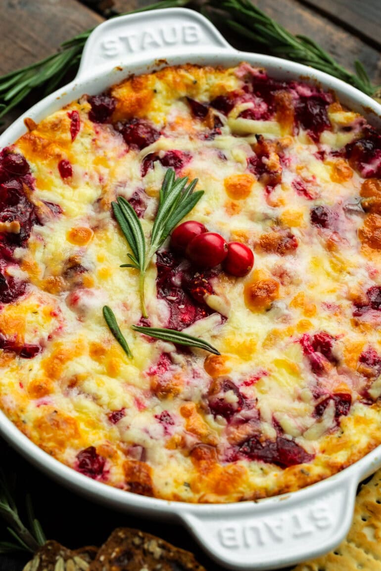 a circular baking dish full of cranberry cream cheese dip topped with melted cheese, a sprig of fresh rosemary and three whole cranberries