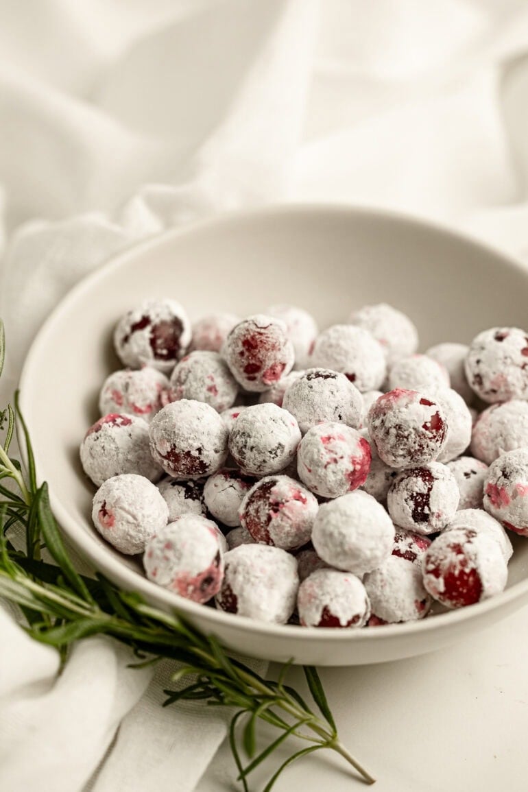 a small bowl of sugar coated whole cranberries with a sprig of fresh rosemary laying next to it.