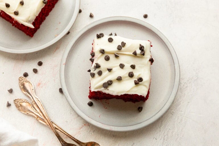 a slice of red velvet cake topped with cream cheese frosting and mini chocolate chips on a small white dessert plate