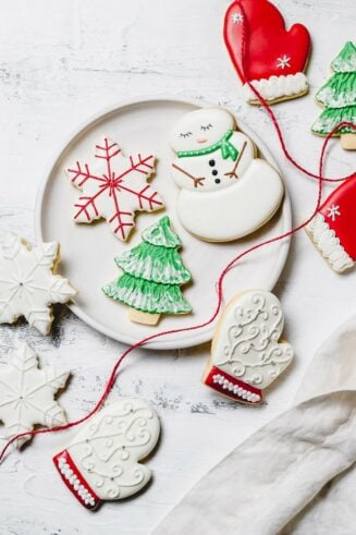 A plate of Christmas sugar cookies decorated with royal icing. There is a snowman with green scarf, a snowflake with red accents, a green tree with white snow, a white mitten with red trim, and white snowflakes