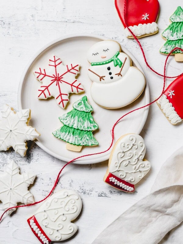 A plate of Christmas sugar cookies decorated with royal icing. There is a snowman with green scarf, a snowflake with red accents, a green tree with white snow, a white mitten with red trim, and white snowflakes