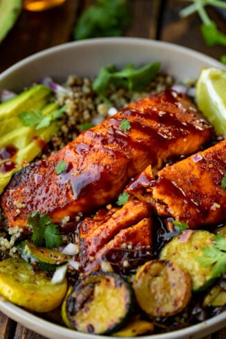 a photo of two salmon fillets glazed with bbq sauce sitting on a plate surrounded by grilled zucchini slices, lime wedges and sliced avocado