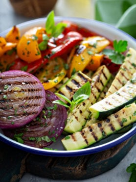 A serving dish of grilled veggies. The zucchini wedges, sliced red onions, and red and yellow bell peppers have perfect grill marks. and the veggies have a few parsley leaves scattered on top.