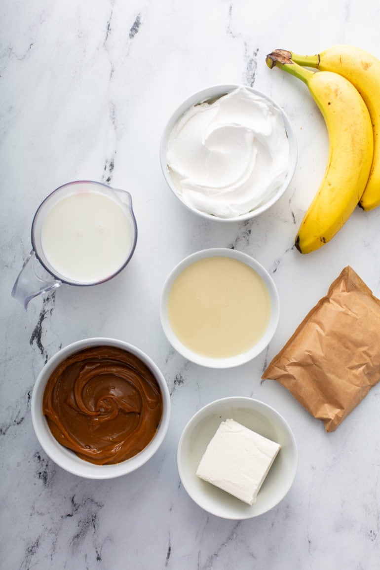 Ingredients banoffee pie. There are containers of dulce de leche, pudding, milk, cream, sugar, cream cheese and bananas.