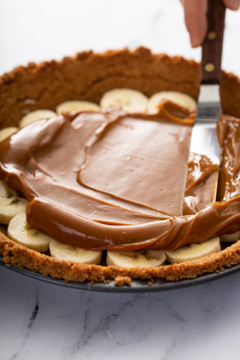 A graham cracker crust in a pie dish. There are sliced bananas in the bottom of the crust and dulce de leche is being spread over the top of the bananas.