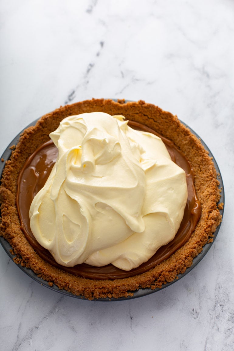 A graham cracker crust filled with dulce de leche and banana cream filling that has been added, but not yet spread all over the dulce de leche.