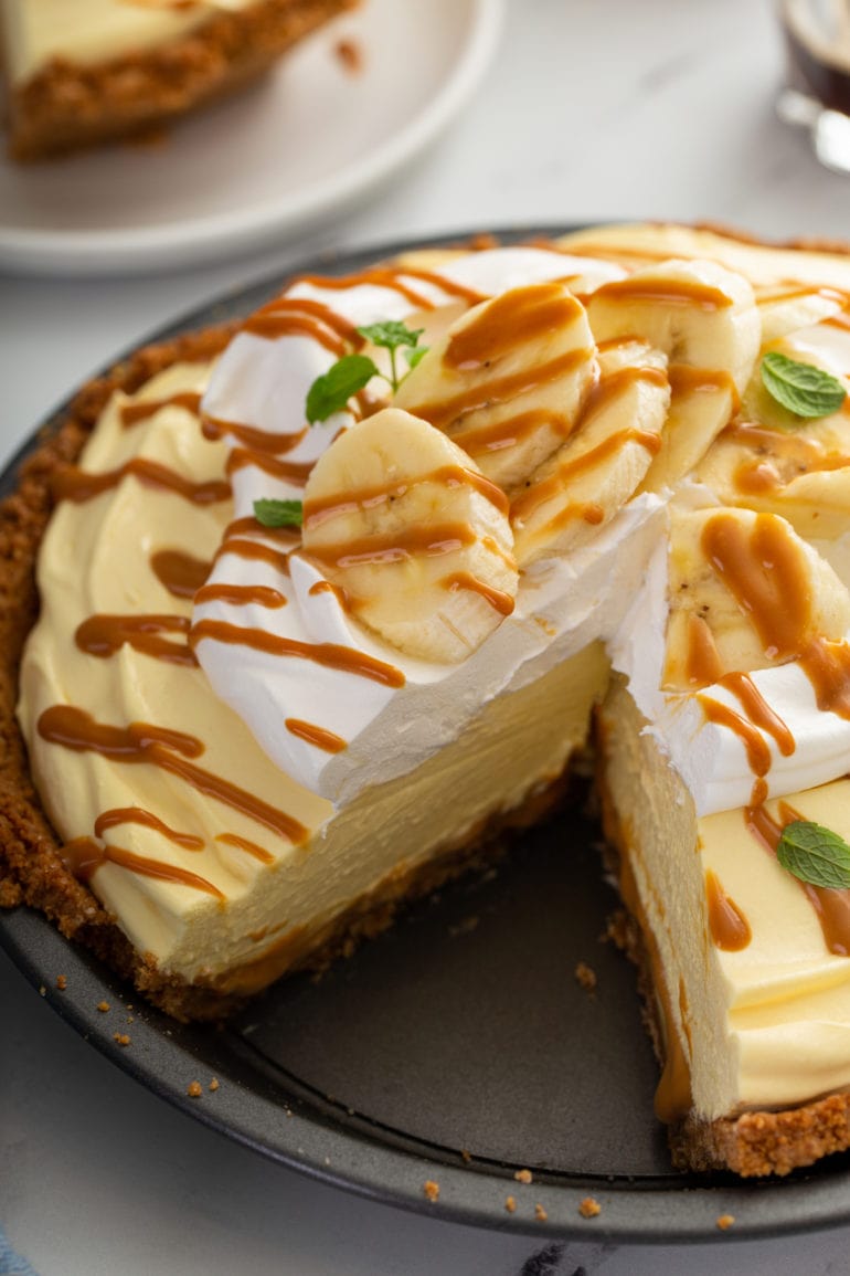 A banoffee pie with whipped cream and sliced bananas on top. The whole pie is drizzled with dulce de leche and a slice has been cut from the pie.