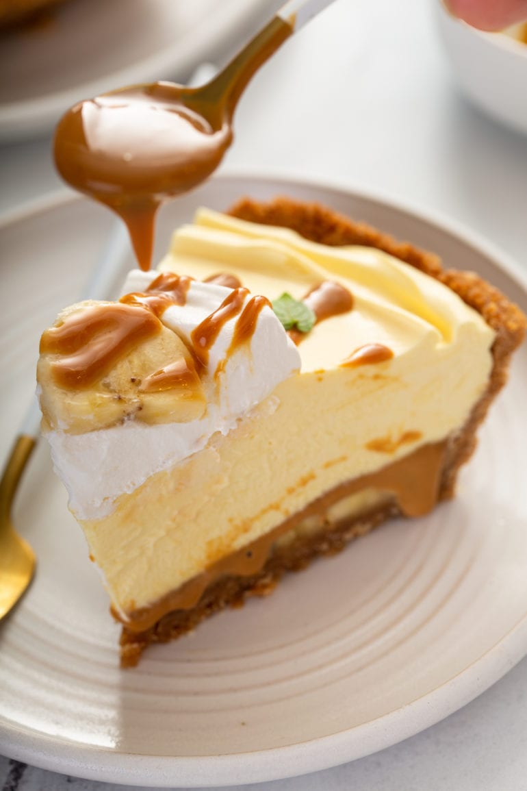 A slice of banoffee pie. The pie has a graham cracker crust and is a golden yellow color. There is a dollop of whipped cream on top and dulce de leche is being drizzled over the top of the whipped cream.