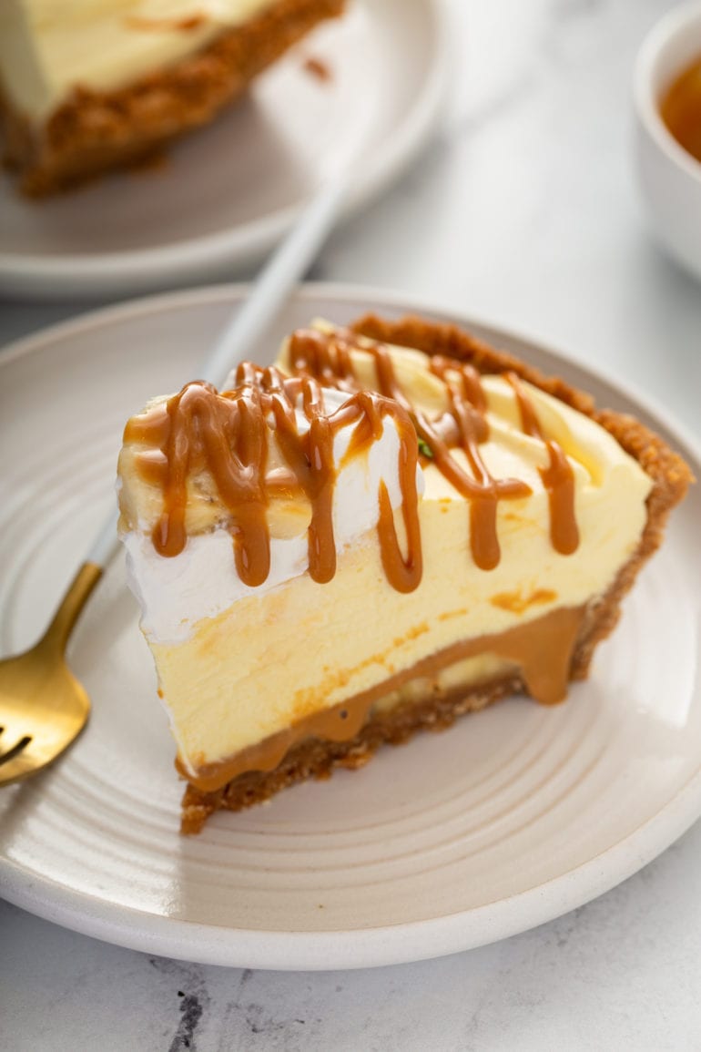 A slice of banoffee pie in a graham cracker crust. there is whipped cream on top and dulce de leche is drizzled over the top of the slice of pie. A fork rests on the plate next to the pie.