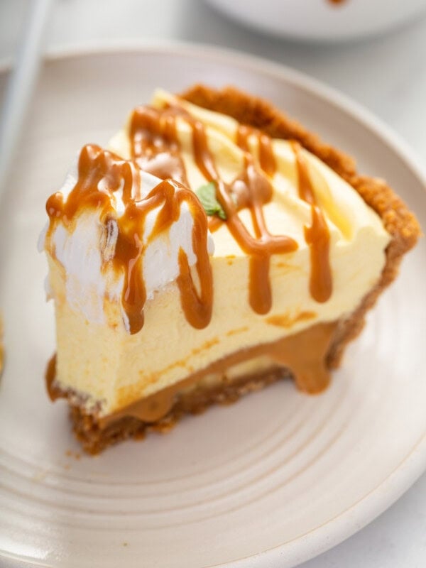A slice of dulce de leche banana cream pie on a white dessert plate. One bite has been taken from the end of the slice. The pie has a dollop of whipped cream on top and then is drizzled with dulce de leche.