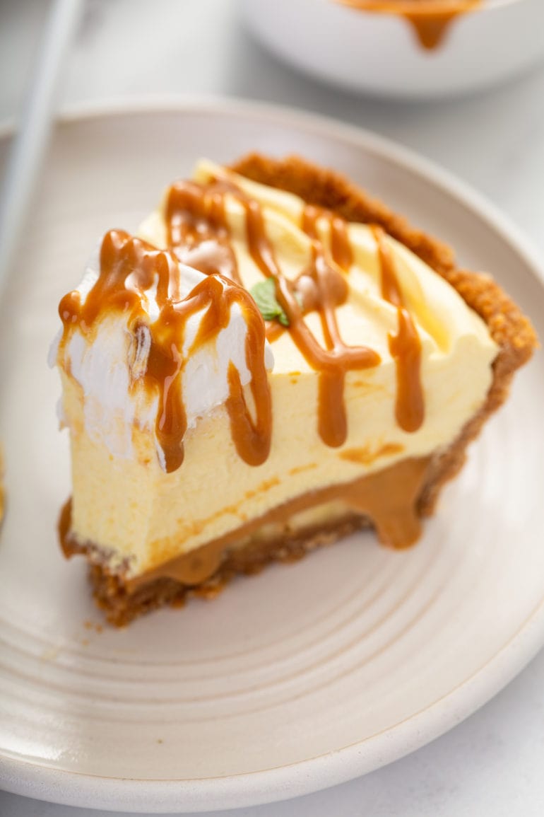 A slice of dulce de leche banana cream pie on a white dessert plate. One bite has been taken from the end of the slice. The pie has a dollop of whipped cream on top and then is drizzled with dulce de leche.