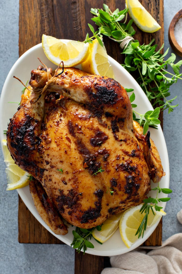 A whole chicken roasted to a golden brown. The chicken is on a white platter and has a few slices of lemon around it and some fresh thyme sprigs next to it.