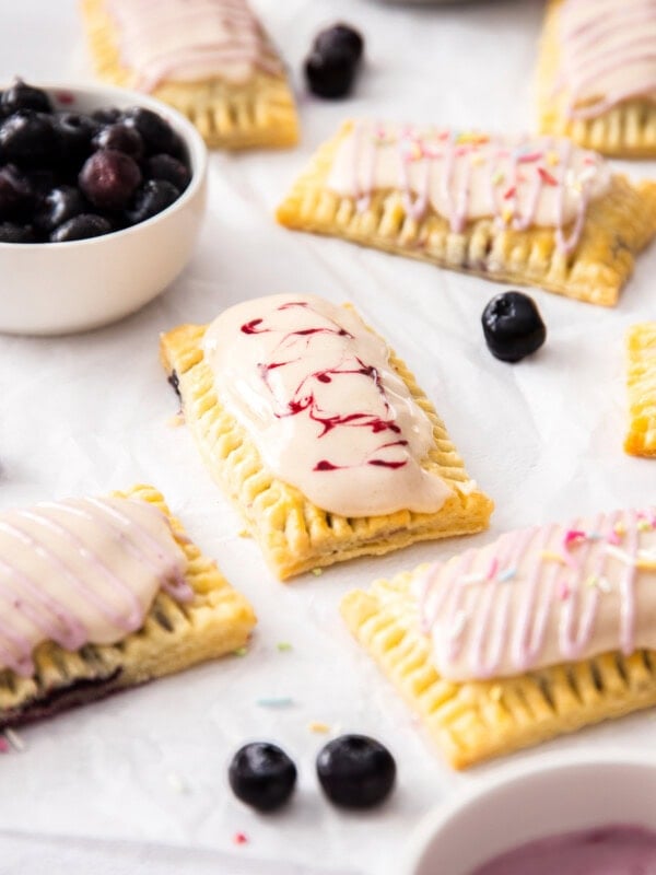 A batch of homemade pop tarts on a sheet of white baking paper. A small white bowl of blueberries.