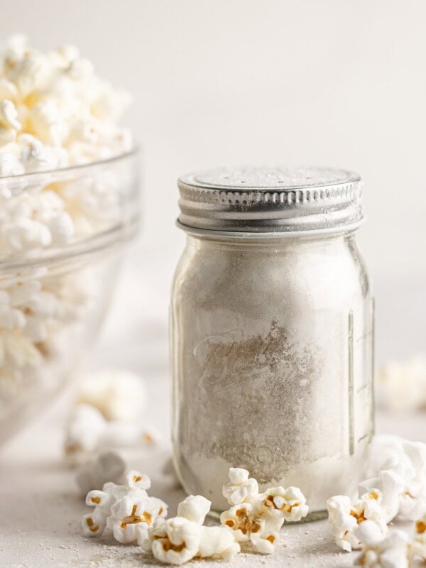 a small glass shaker jar full of caramel popcorn seasoning sitting next to a large glass bowl full of popped popcorn.