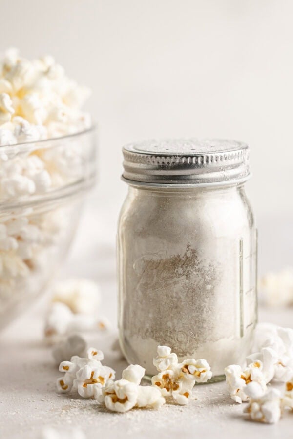 a small glass shaker jar full of caramel popcorn seasoning sitting next to a large glass bowl full of popped popcorn.