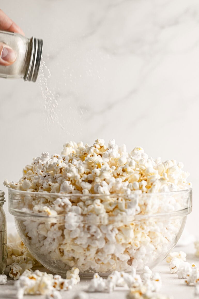 caramel popcorn seasoning being shaken from a glass shaker bottle over the top of a bowl full of popped popcorn