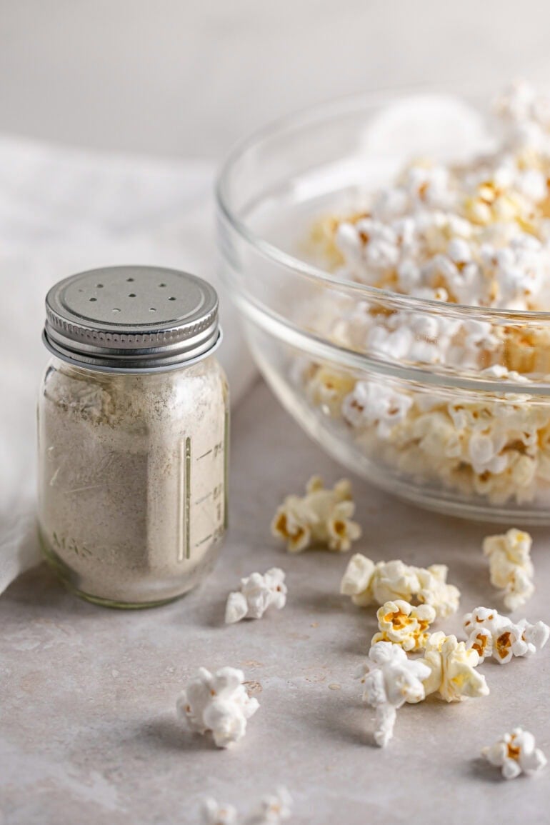 a glass shaker jar of homemade popcorn seasoning sitting next to a glass bowl of popped popcorn