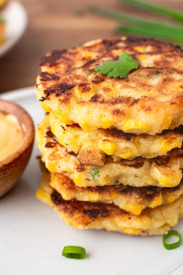 a photo of a stack of crispy golden cheesy corn fritters with a small bowl of dip next to it