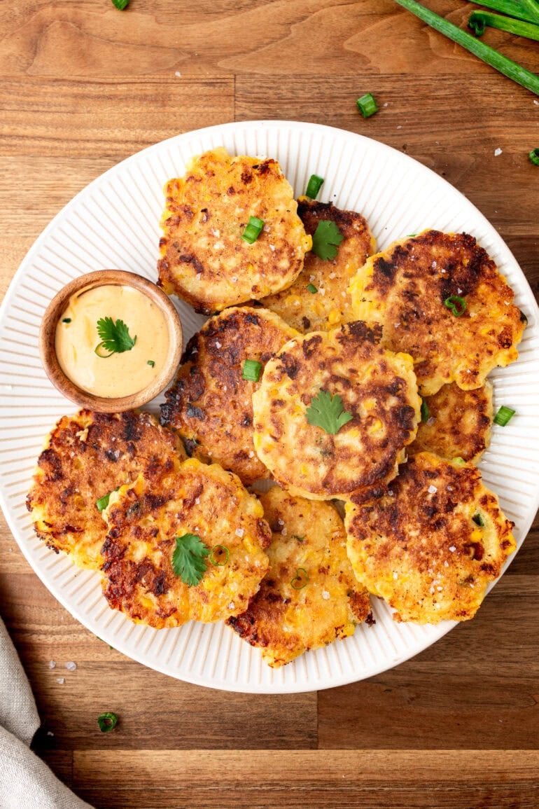 a photo of several golden crispy corn fritters sitting on a large white plate with a small cup of dip on the side