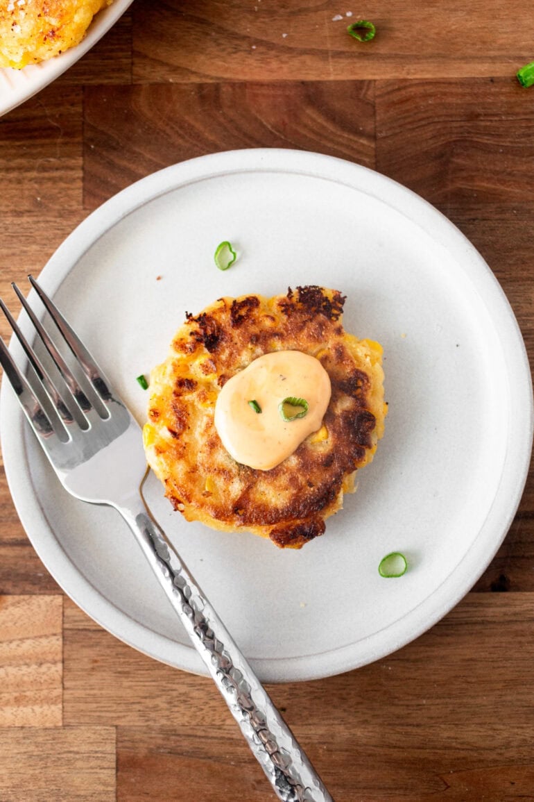 a photo of a single corn fritter sitting on a small plate topped with a dollop of dip on top and a fork sitting on the side