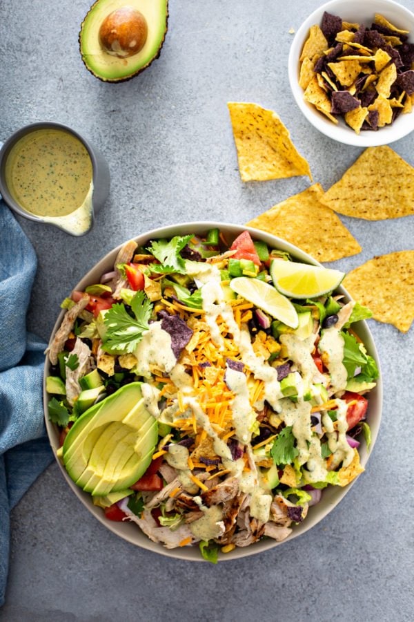 Chicken taco salad in a large serving bowl. There is chicken, tomatoes, corn, beans, red onions, peppers and cheese. A sliced avocado and 2 lime wedges are on top. There are chips and a container of dressing are next to the salad.