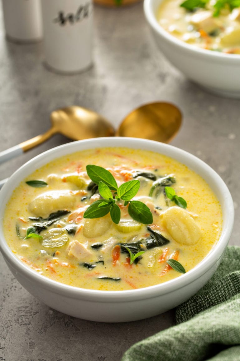 A bowl of copycat gnocchi soup. There are shredded carrots, gnocchi, celery, and chicken in a creamy soup. Two soup spoons are next to the bowl of soup and a sprig fresh herbs tops the soup.
