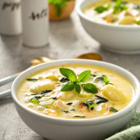 Two bowls of copycat Olive Garden chicken gnocchi soup. The soup has shredded carrots, gnocchi, celery and spinach in a creamy soup. Fresh herbs are sprinkled on top, and two soup spoons, salt and pepper shakers, and a green napkin are on the table next to the soup.