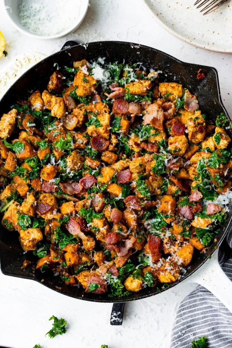 a large cast iron skillet full of golden chunks of chicken breast mixed with sauteed chopped kale, cubed sweet potatoes, bacon bits and sprinkled with parmesan cheese