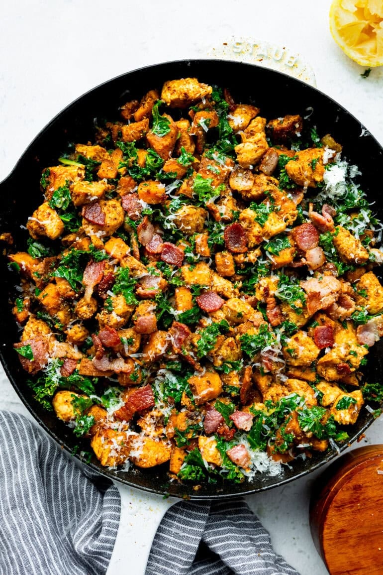 a cast iron skillet full of golden chunks of chicken mixed with bacon crumbles, kale and sweet potatoes