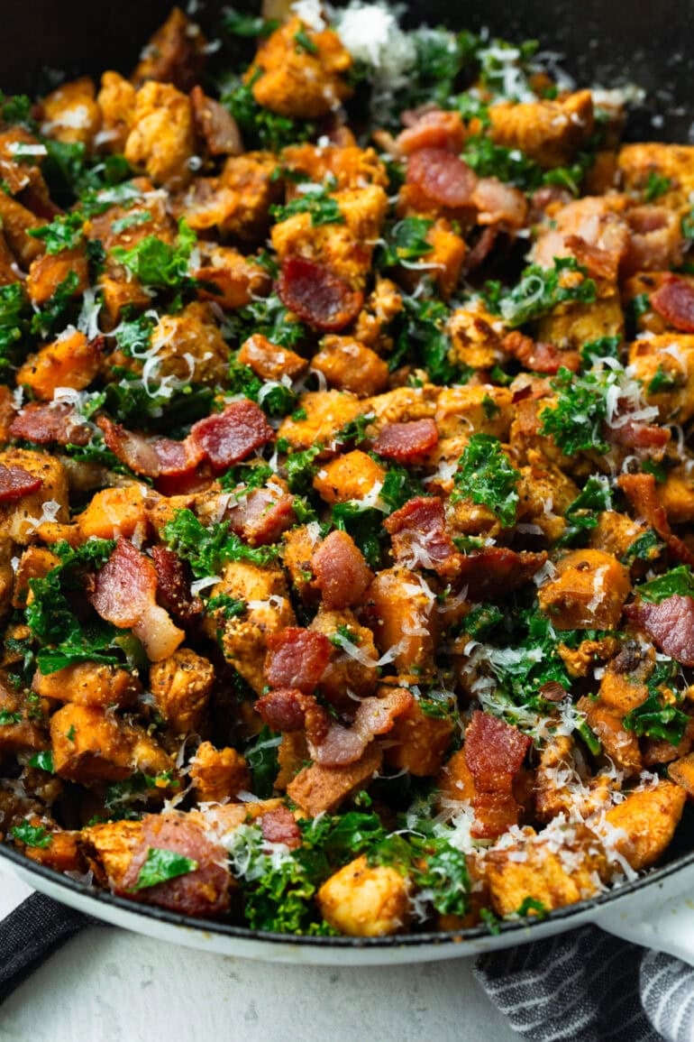 a cast iron skillet with cooked chicken chunks, cubed sweet potatoes, sauteed kale, bacon bits and parmesan cheese