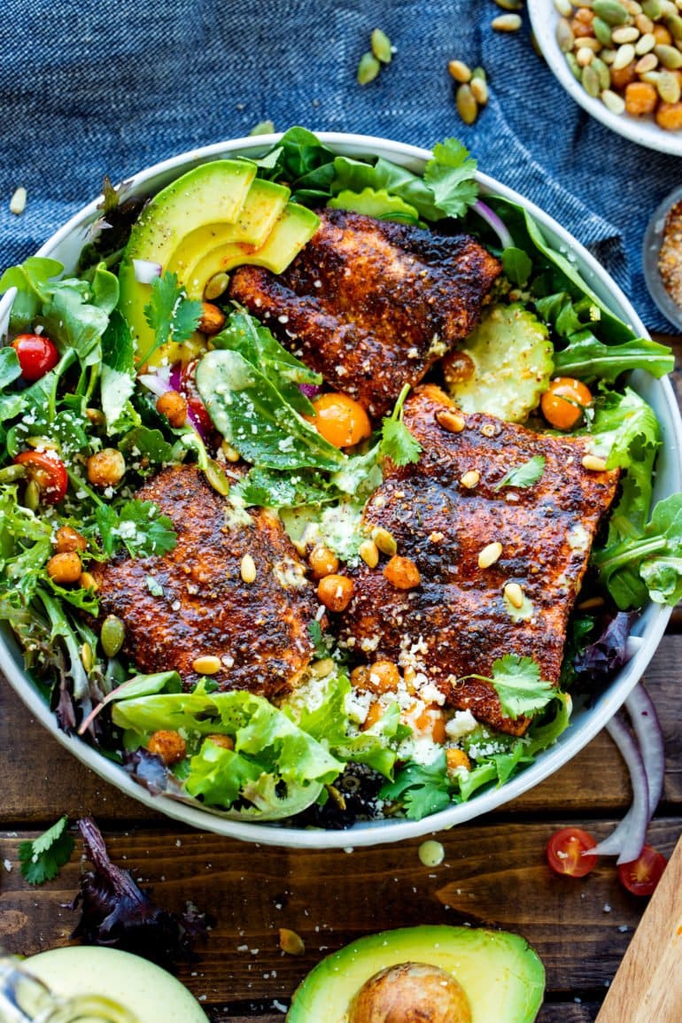 a photo of a bright arugula cucumber salad in a serving bowl topped with three seasoned baked salmon filets, roasted chickpeas and toasted pine nuts and pepitas.