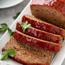 A tray holding meatloaf that has four slices cut, sprinkled with parsley leaves, with a side dish of green beans.