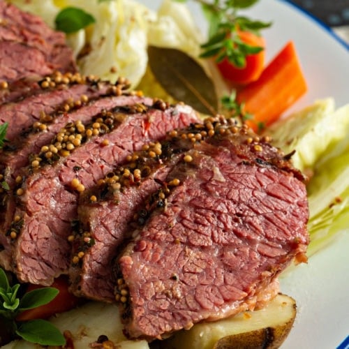 Slice corned beef resting on top of cabbage, potatoes and carrots with sprigs of herbs on the side.