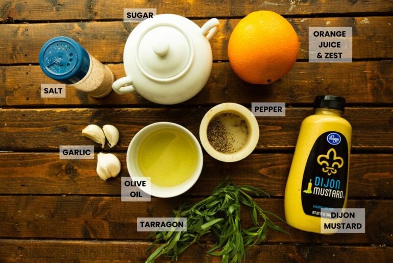 Plain de ingredientes clave: dientes de ajo, aceite de oliva, pimienta, mostaza Dijon, estragón fresco, una naranja y una tetera, dispuesta en una superficie rústica de madera.