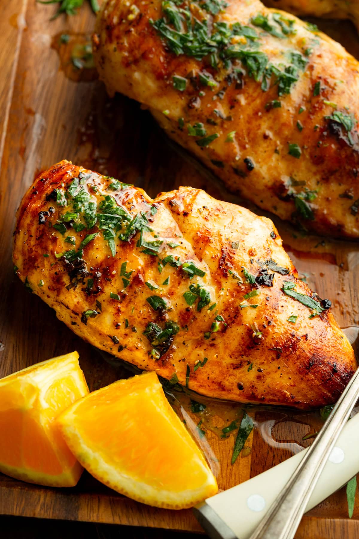 Múltiples pechugas de pollo a la parrilla en una bandeja de madera con rodajas de naranja, brillantes con marinadas de cítricos y hierbas frescas.