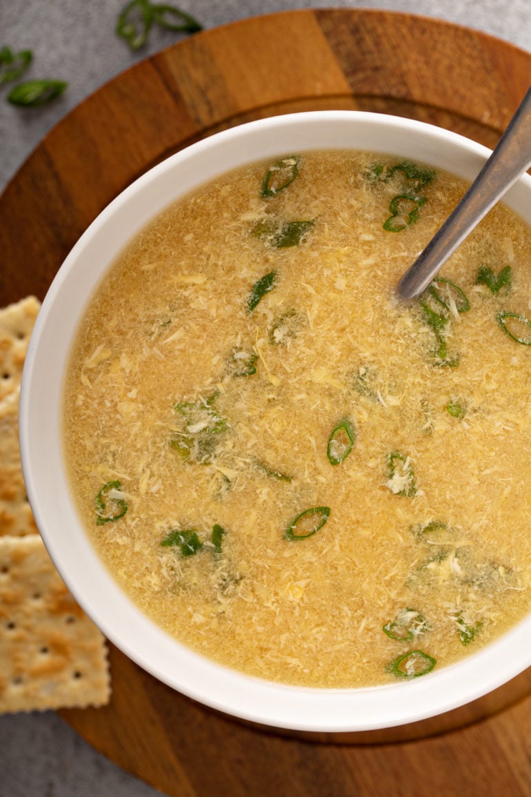 A close-up of a bowl of egg drop soup with sliced onions on top and saltine crackers in the background.
