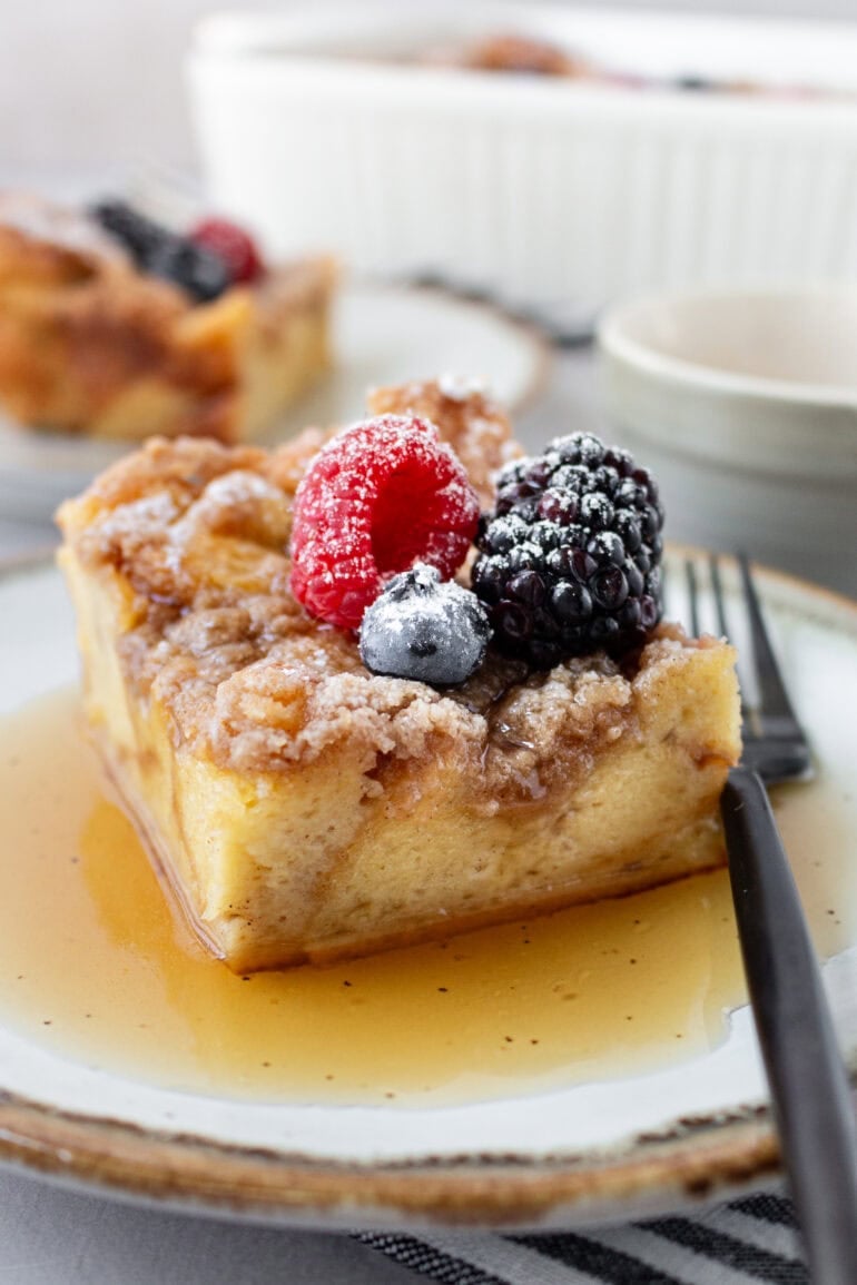 A photo of a piece of French toast casserole in maple syrup on a plate with a fork next to it and topped with fresh berries