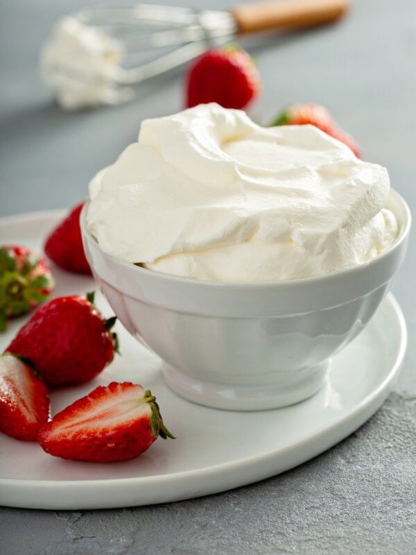 A bowl of whipped cream sitting on a plate with a few sliced and whole strawberries. There is a whisk and two strawberries in the background.