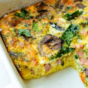 a baking dish of a baked egg, spinach and sauteed mushroom casserole with a serving missing so you can see the cross-section of the casserole