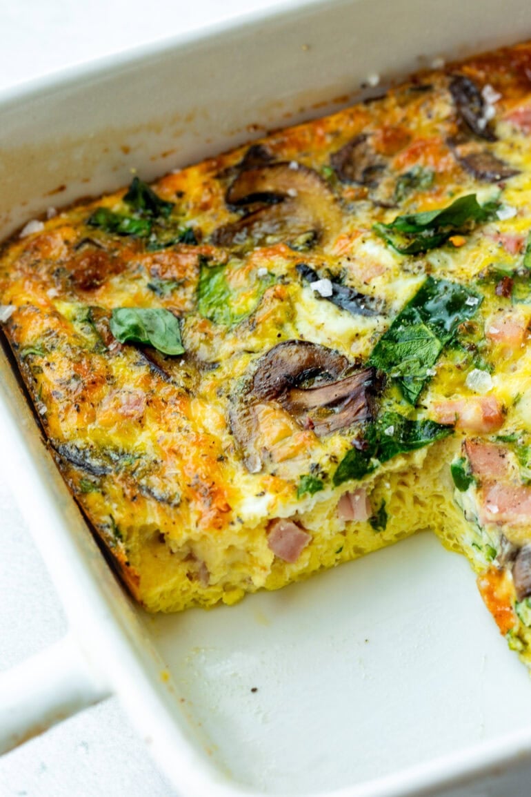 a baking dish of a baked egg, spinach and sauteed mushroom casserole with a serving missing so you can see the cross-section of the casserole