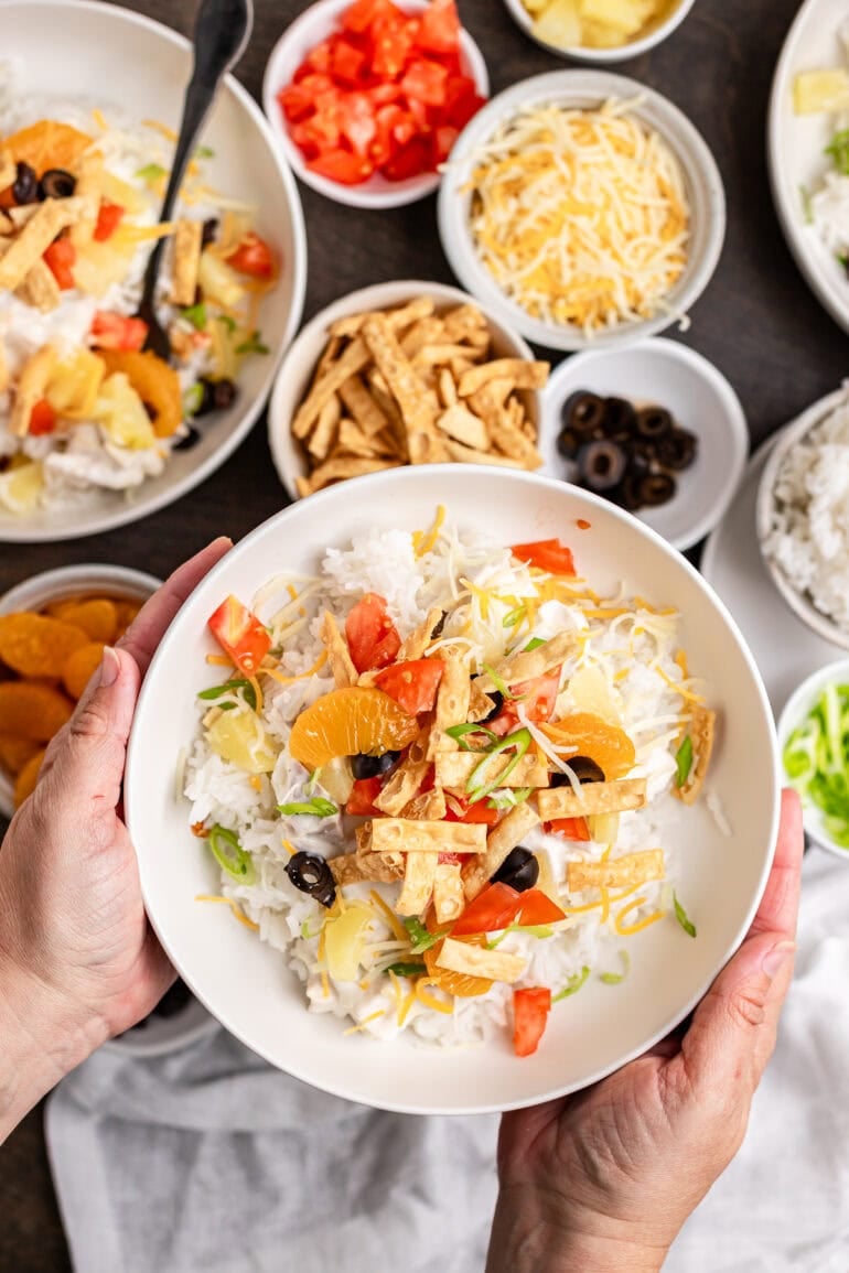 A white bowl filled with Hawaiian Haystacks topped with pineapple chunks, cheddar cheese, green onions, and fresh tomatoes.