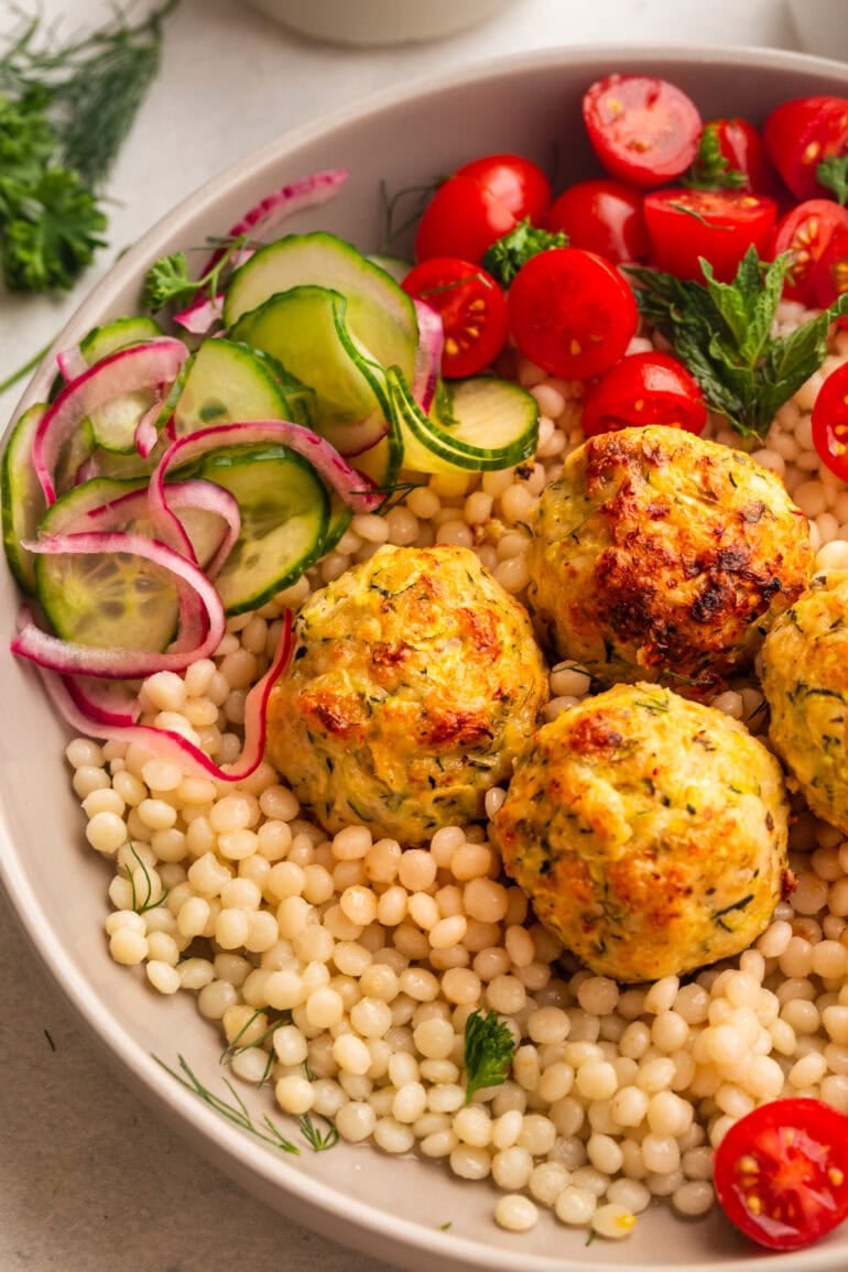 golden chicken meatballs on a bed of pearl couscous with cherry tomatoes and a cucumber salad in the background
