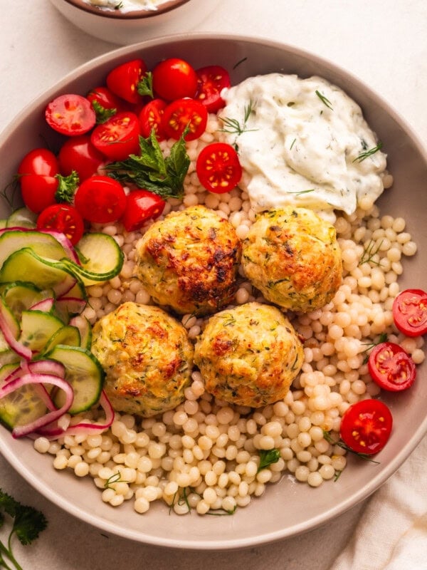 a plate of pearl cous cous topped with chicken meatballs with a side of cherry tomatoes and tzatziki