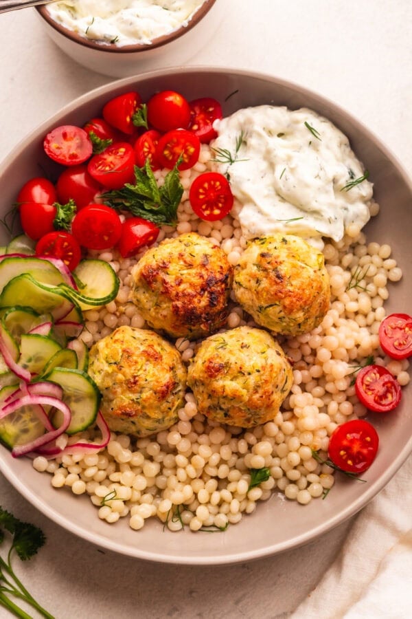 a plate of pearl cous cous topped with chicken meatballs with a side of cherry tomatoes and tzatziki
