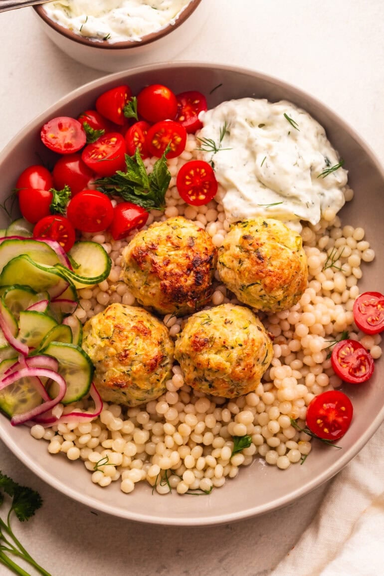 a plate of pearl cous cous topped with chicken meatballs with a side of cherry tomatoes and tzatziki