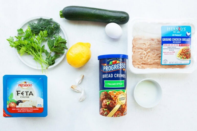 all the ingredients for herb greek chicken meatballs including ground chicken, an egg, breadcrumbs, lemon, fresh herbs, zucchini, garlic and feta cheese