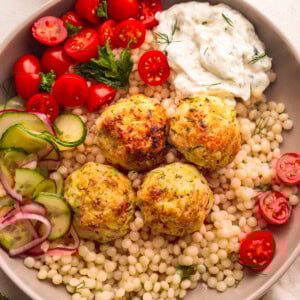 a plate of pearl cous cous topped with chicken meatballs with a side of cherry tomatoes and tzatziki