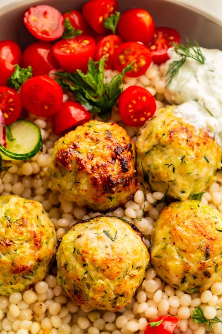 several golden baked herb Greek chicken meatballs on top of a bed of pearl couscous and halved cherry tomatoes in the background