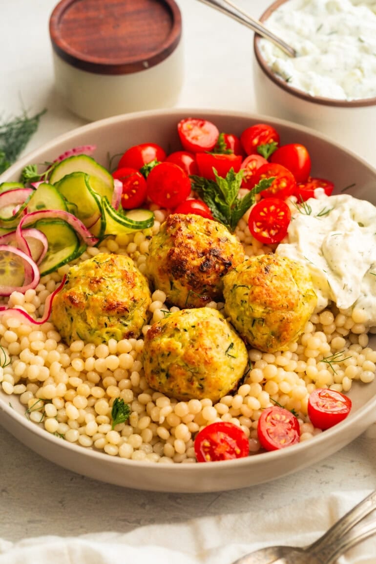 a bowl of pearl cous cous topped with greek chicken meatballs with tzatziki sauce, cherry tomatoes and a cucumber salad
