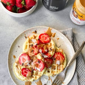 a photo of three small circular waffles topped with sliced strawberries on a white plate with a fork sitting on the side of the plate